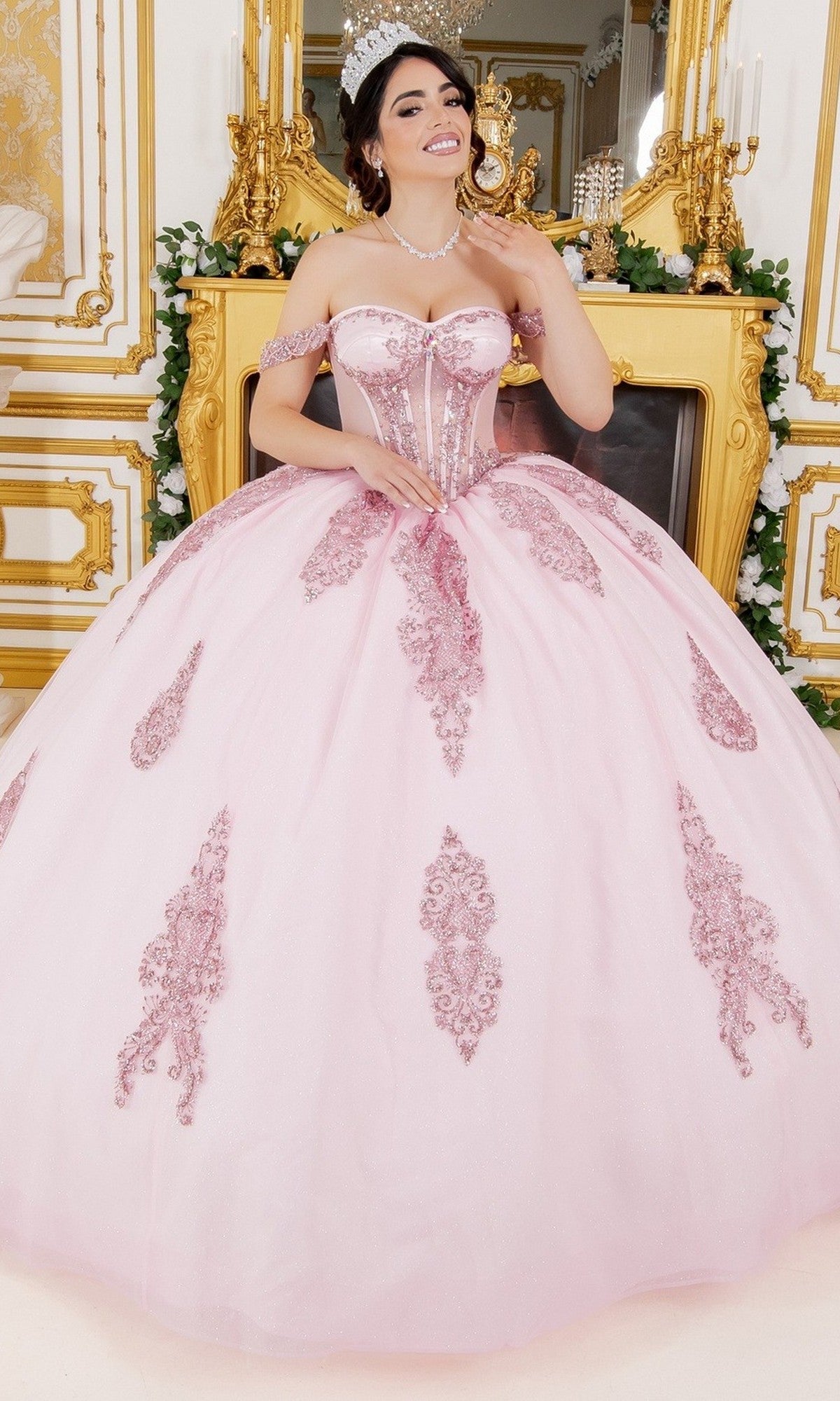 A young woman, wearing the Quinceanera Dress 8190J by Cinderella Couture in light pink with ornate embroidery, poses in front of a gold-trimmed fireplace, smiling and accessorized with a tiara.